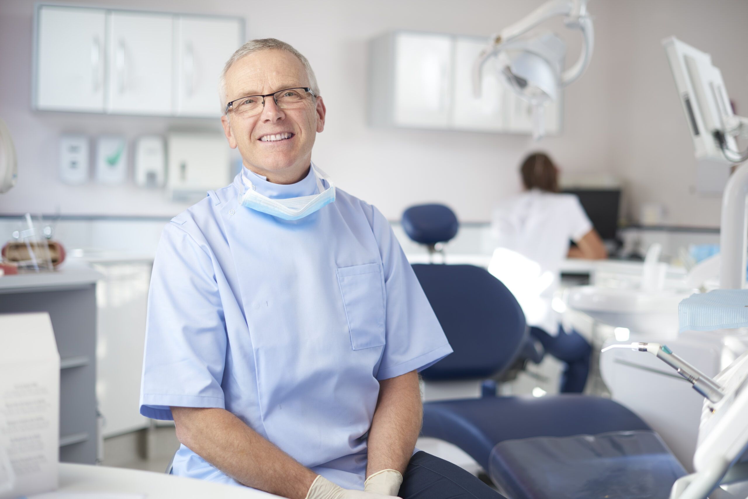 A mature male dentist is looking proudly to camera from his dentist's office. In the background a dental nurse is prepping for the next patient.